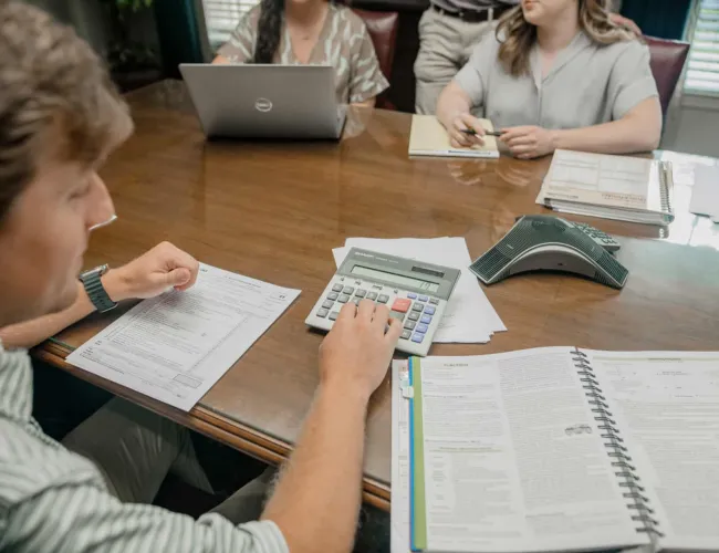 Group of professionals at wooden table using calculator, laptop, and documents during office meeting.