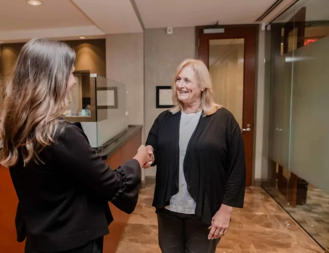 Two women shaking hands in a modern office reception area, smiling and greeting each other warmly.