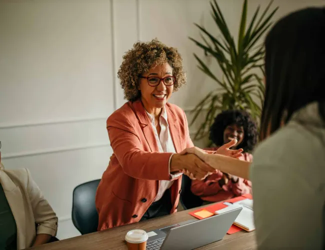 Smiling woman in a pink blazer shaking hands with another person during a business meeting at a desk.