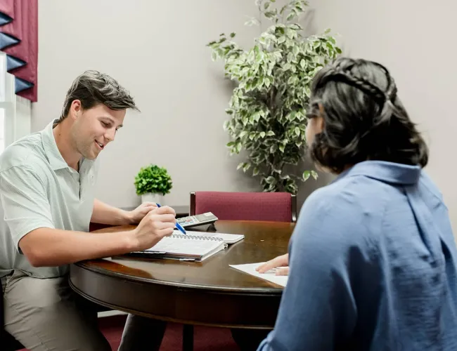 Two people sitting at a round table discussing notes with a calculator and plant in background