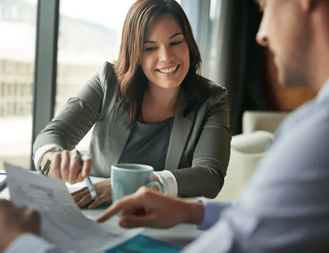 Businesswoman smiling and discussing documents with a male colleague in an office setting over coffee.
