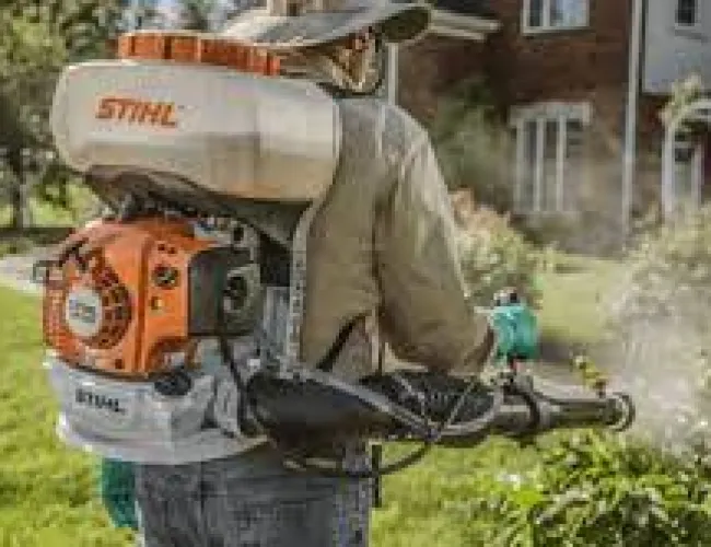 Person using a STIHL backpack sprayer to spray plants in a garden near a house on a sunny day