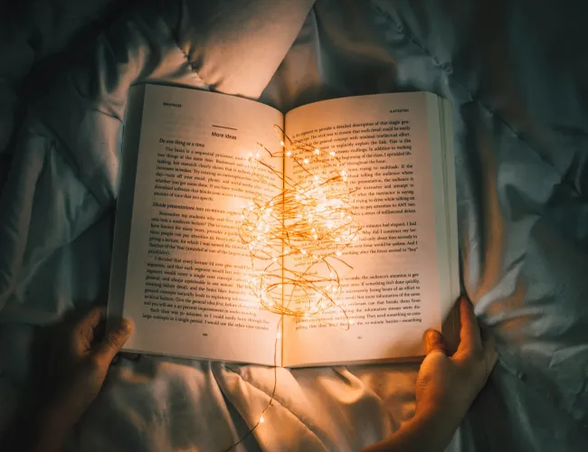 Open book held over a bed with warm fairy lights illuminating the pages in a cozy setting