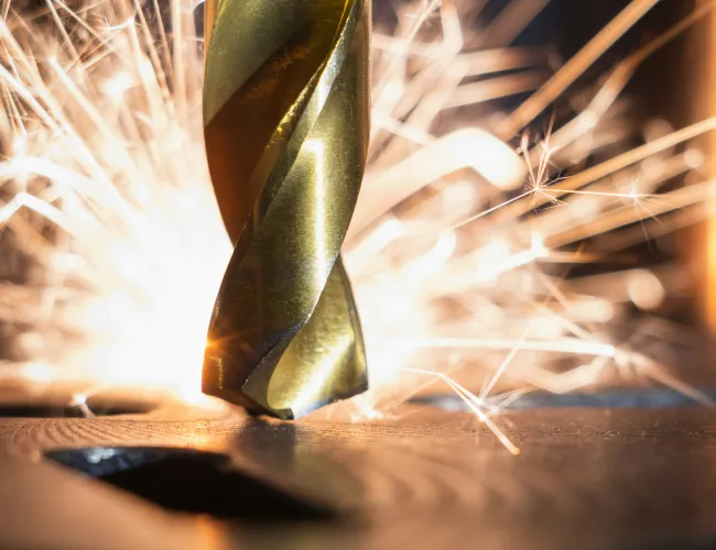 Close-up of a gold drill bit creating sparks while drilling into metal with a blurred dark background.