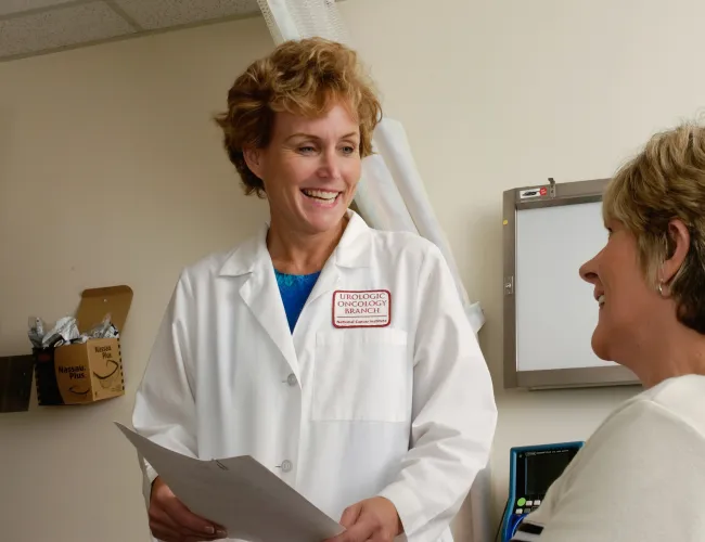 Female doctor in white coat discussing medical report with smiling female patient in clinic room.