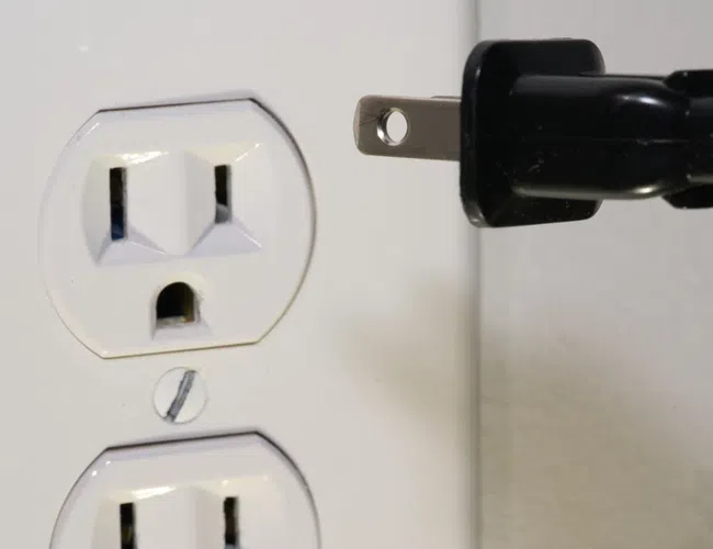 Black electrical plug about to be inserted into a white wall power outlet with two sockets.