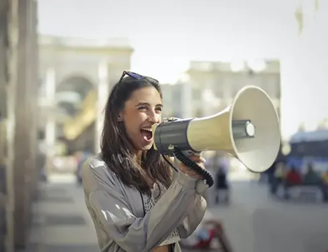 Woman using a megaphone outdoors with a city background, appearing excited and expressive.