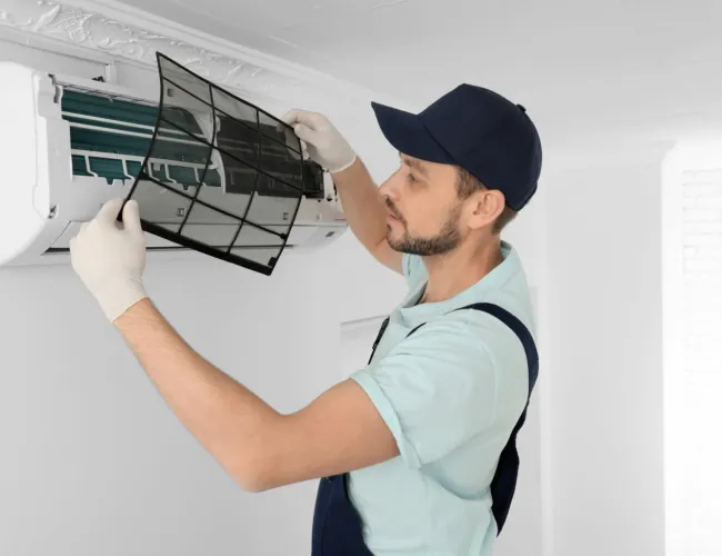 Technician wearing gloves and cap cleaning or replacing air conditioner filter indoors in a bright room