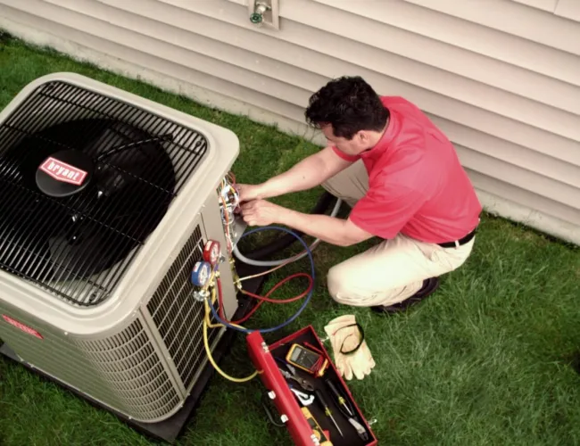 Technician in red shirt repairing a Bryant air conditioning unit outside a home on green grass near siding.