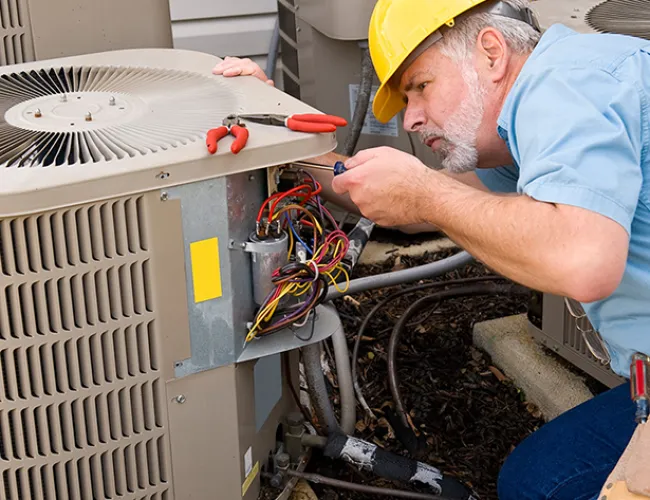Technician in a yellow hard hat repairing an outdoor air conditioning unit with tools and exposed wiring