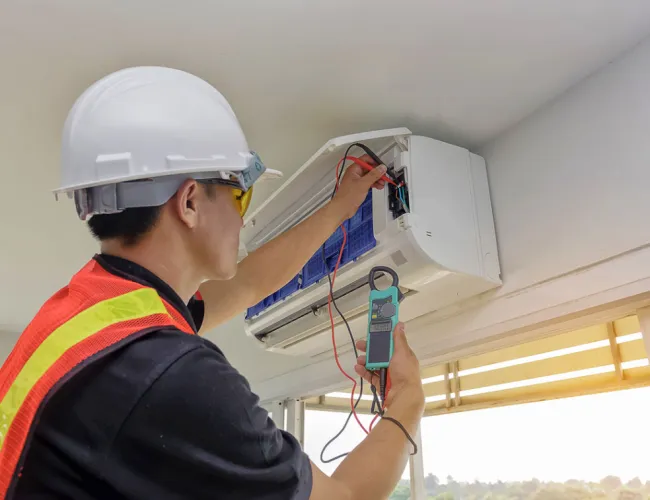 Technician wearing safety gear repairing and testing a wall-mounted air conditioning unit indoors with multimeter.