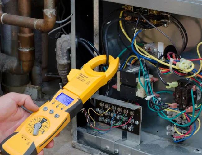 Hand holding yellow clamp meter measuring current on electrical control panel wiring.