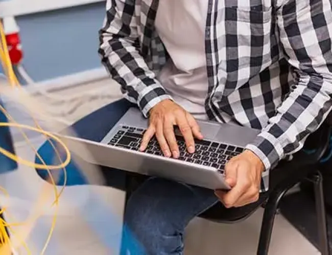 Technician working on a laptop in a network server room with yellow Ethernet cables nearby.
