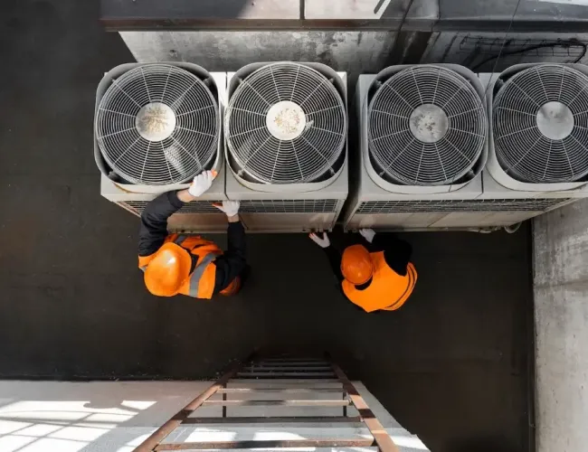 Two workers in orange safety gear inspecting industrial air conditioning units from above in a maintenance area