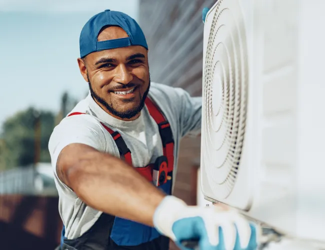 Smiling HVAC technician in blue cap and gloves installing or repairing an air conditioning unit outdoors.