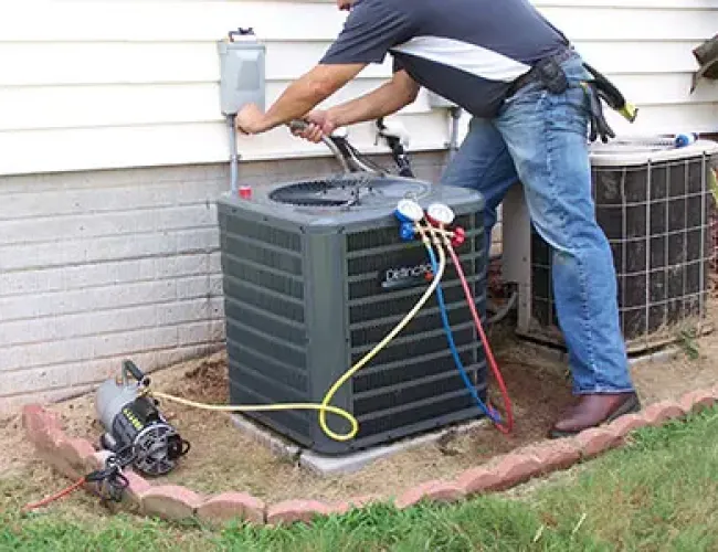 Technician servicing outdoor air conditioning unit with gauges and tools beside a house wall and green grass.
