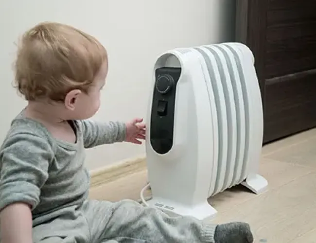 Baby in gray outfit reaching towards a white electric heater on a wooden floor near a dark door.