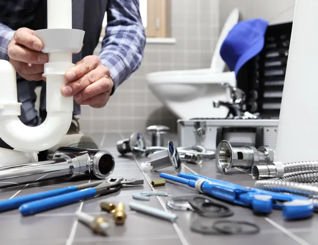 Plumber fitting a white PVC pipe under a bathroom sink with various plumbing tools scattered on the floor.
