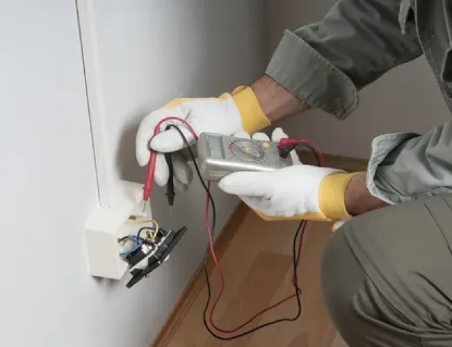 Electrician wearing gloves testing a wall outlet with a multimeter in a residential setting.