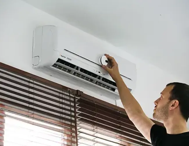 Man adjusting settings on a white wall-mounted air conditioner above window blinds in a bright room