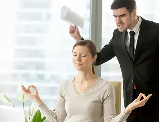 Calm woman meditating at desk while angry man in suit yells and waves paper in office.