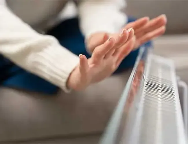 Person with outstretched hands warming near an electric heater indoors, wearing a white sweater.