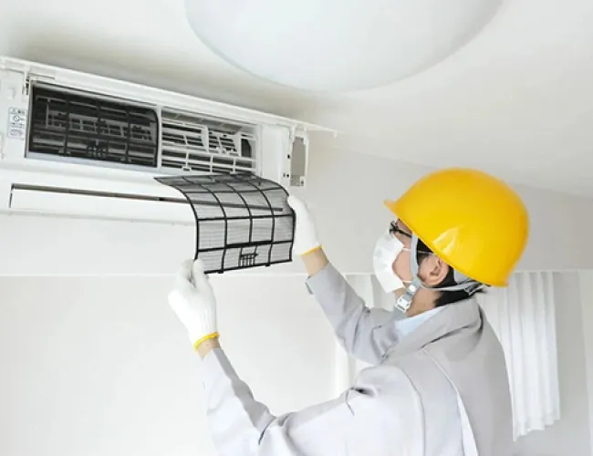 Technician in yellow helmet and mask cleaning or replacing air conditioner filter indoors.