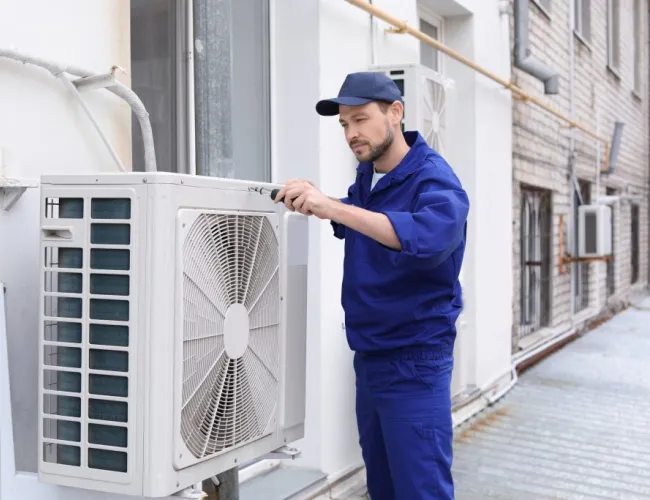 Technician in blue uniform repairing outdoor air conditioning unit on building exterior wall.