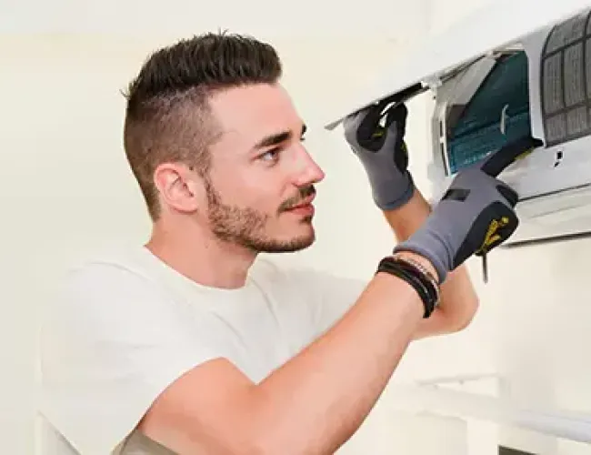 Technician wearing gloves inspecting and repairing a wall-mounted air conditioner unit indoors.