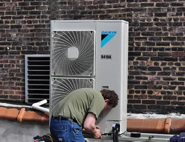 Technician installing or repairing Daikin R410A air conditioning unit on rooftop next to brick wall.