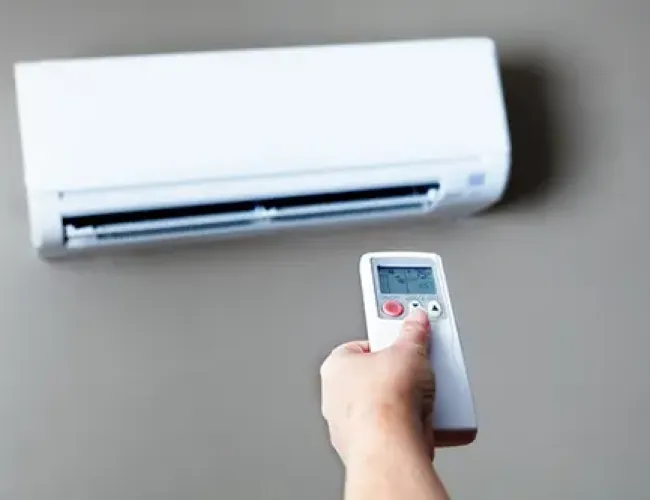 Hand holding remote control pointing at wall-mounted air conditioner unit for temperature adjustment.
