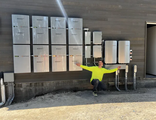 Man in neon yellow shirt kneeling by multiple Enphase electrical panels mounted on a brown exterior wall outdoors.