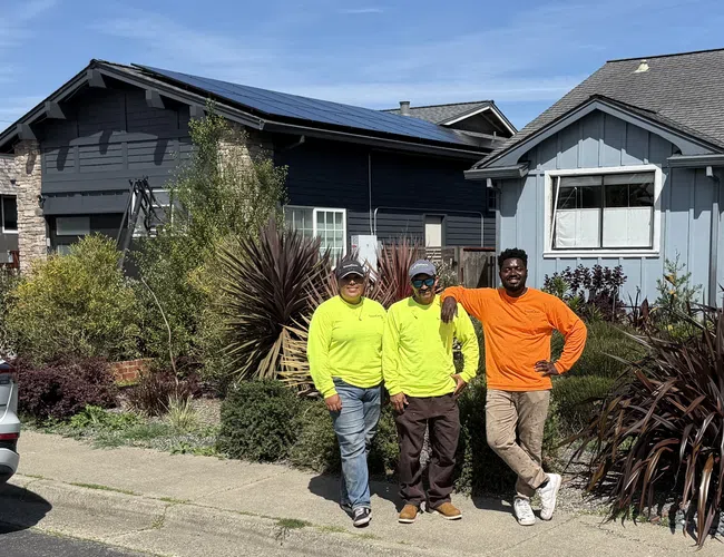 Three workers in bright shirts posing in front of a house with solar panels under clear blue sky