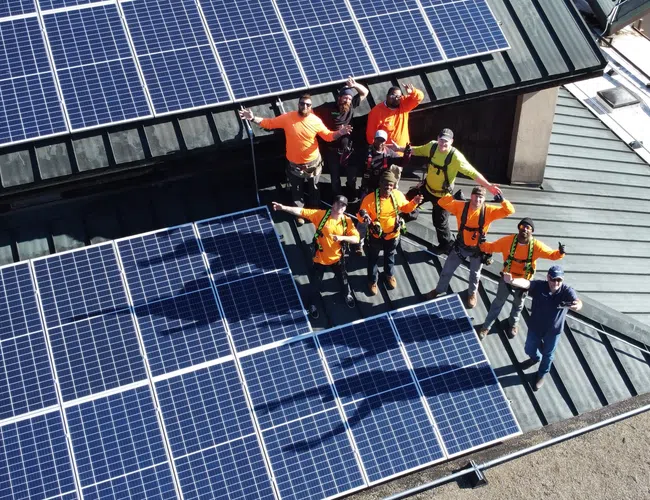 Group of workers in safety gear standing on roof with installed solar panels, waving at the camera.