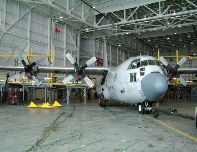 Four-engine propeller aircraft inside a spacious maintenance hangar with equipment and scaffolding around.