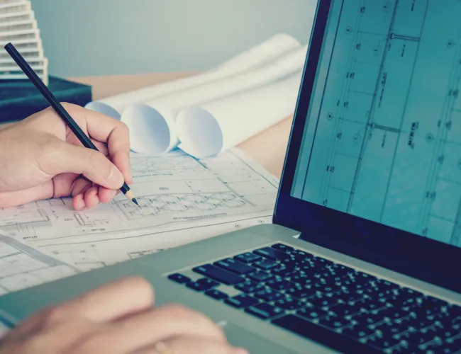 Architect working on building blueprints with pencil and laptop displaying architectural plans.
