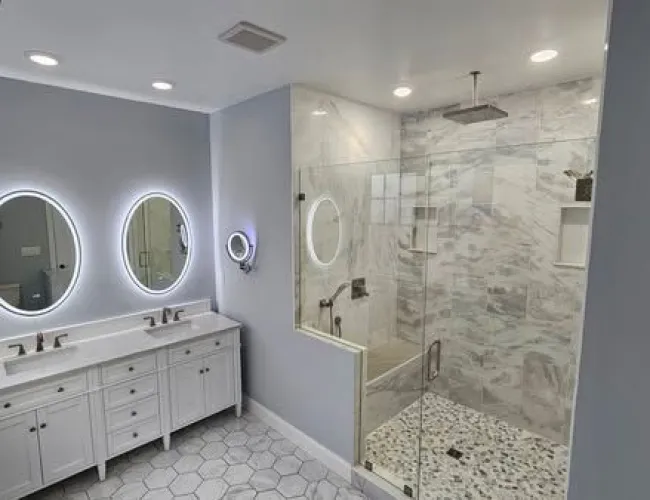 Modern bathroom with double white vanity, illuminated round mirrors, and glass-enclosed rain shower with marble accents
