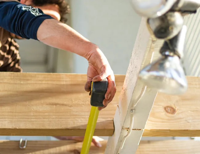 Person using a yellow tape measure to measure a wooden plank in a woodworking or construction setting.