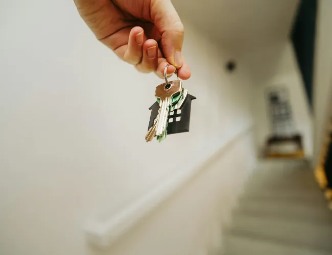Hand holding house-shaped keychain with keys in a blurred staircase background