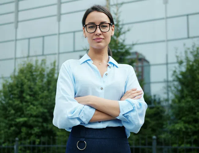 Confident young woman with glasses wearing a blue shirt and dark skirt standing with arms crossed outdoors.