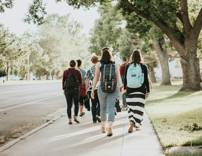 Group of people walking on a tree-lined sidewalk with backpacks on a sunny day.