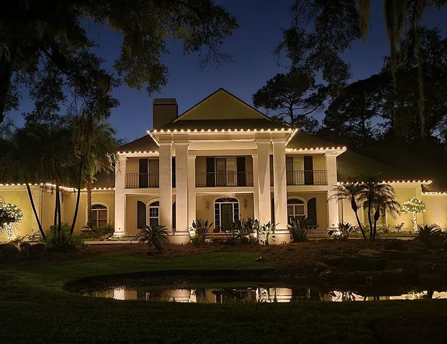 Large illuminated mansion with columns and palm trees at night reflected in a small pond.