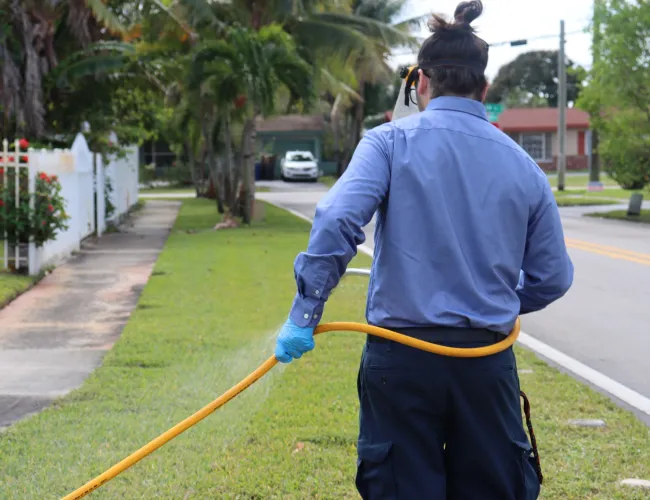 Pest control technician in blue uniform spraying outside a house lawn with safety gloves and goggles.