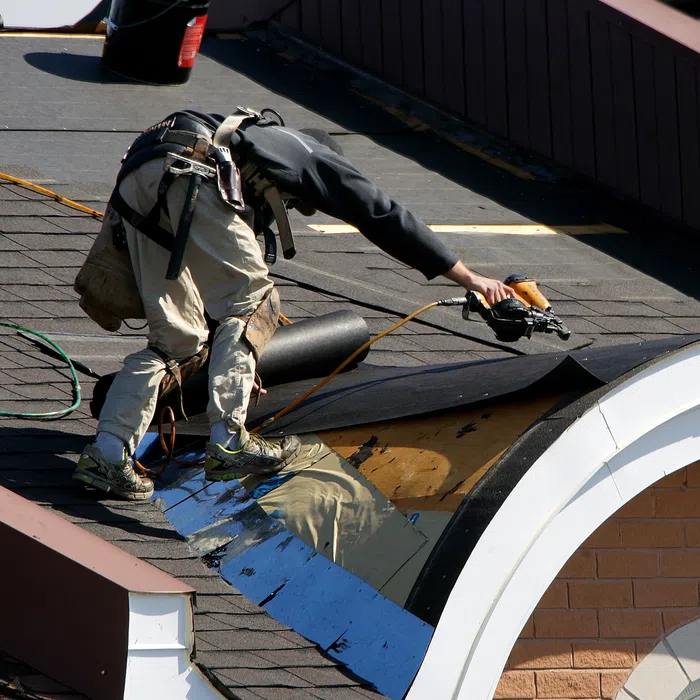 A worker installs shingles on a steep roof, showcasing construction tools and weatherproofing materials.