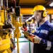 Industrial worker in safety gear inspecting machinery and taking notes on a tablet in a factory setting