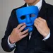 Person in suit holding a blue mask near face against a gray background.