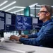 Middle-aged man in glasses working on multiple monitors displaying code in a modern office environment