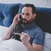 Man with scarf lying in bed checking thermometer while talking on phone, appearing sick or unwell.