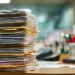 Tall stack of assorted documents and file folders piled on a cluttered office desk with blurred background.