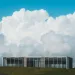 Modern glass building on grassy hill under vast blue sky with large white fluffy clouds.